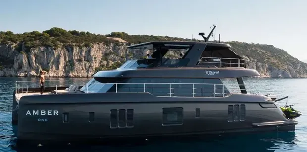 Side view profile of AMBER ONE anchored in calm blue water beside a rocky coastline